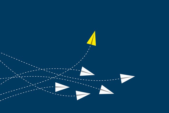 Yellow paper airplane flying upward, away from a cluster of white paper airplanes