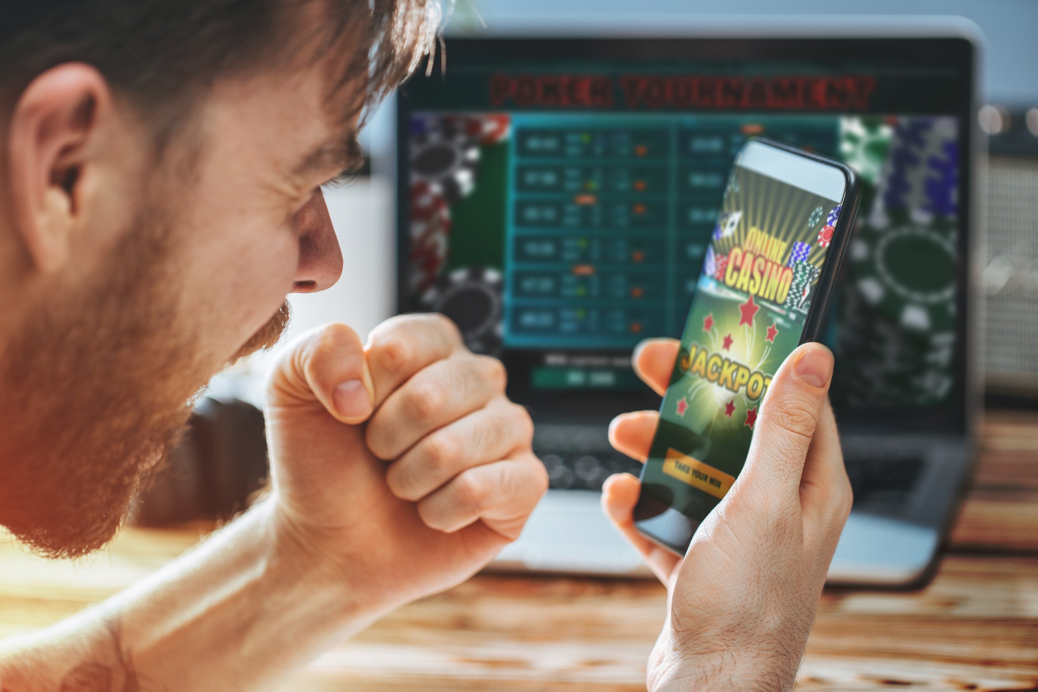 a young man pumps his fist at his phone which is on an online gambling app. 