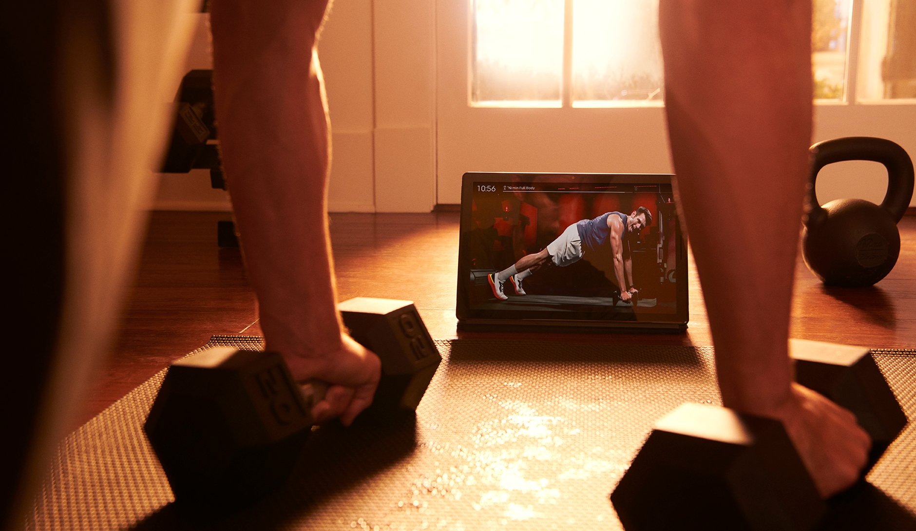 A person does an exercise with two barbells while watching Peloton video content.