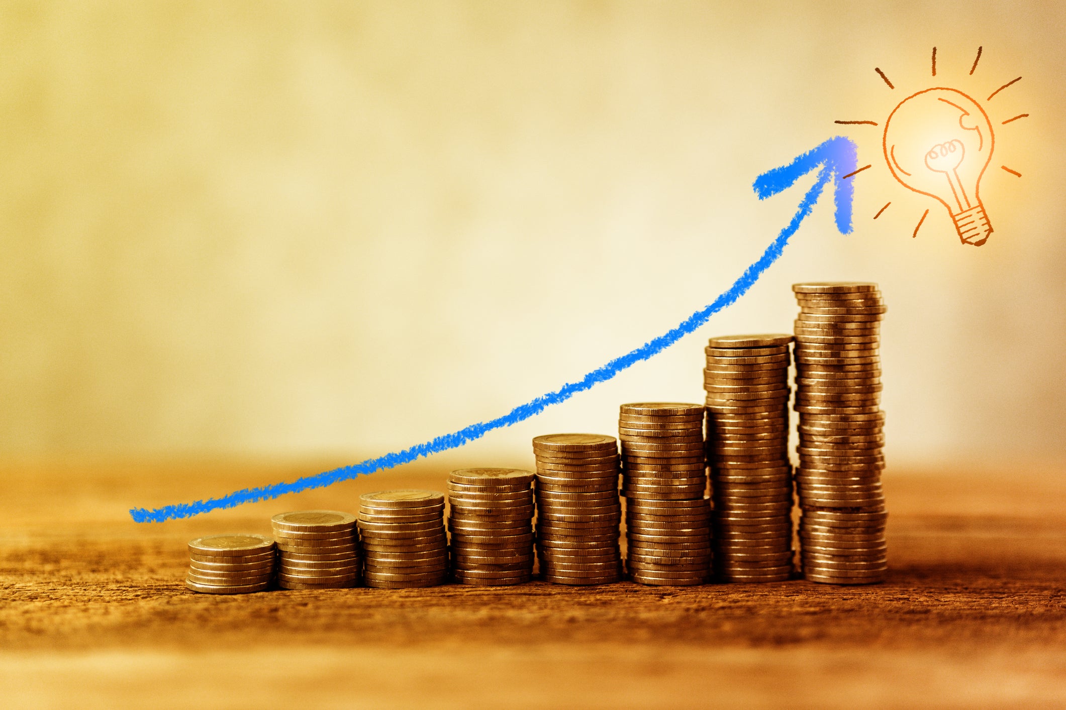 A stack of coins getting progressively higher leading to an illuminated light bulb.