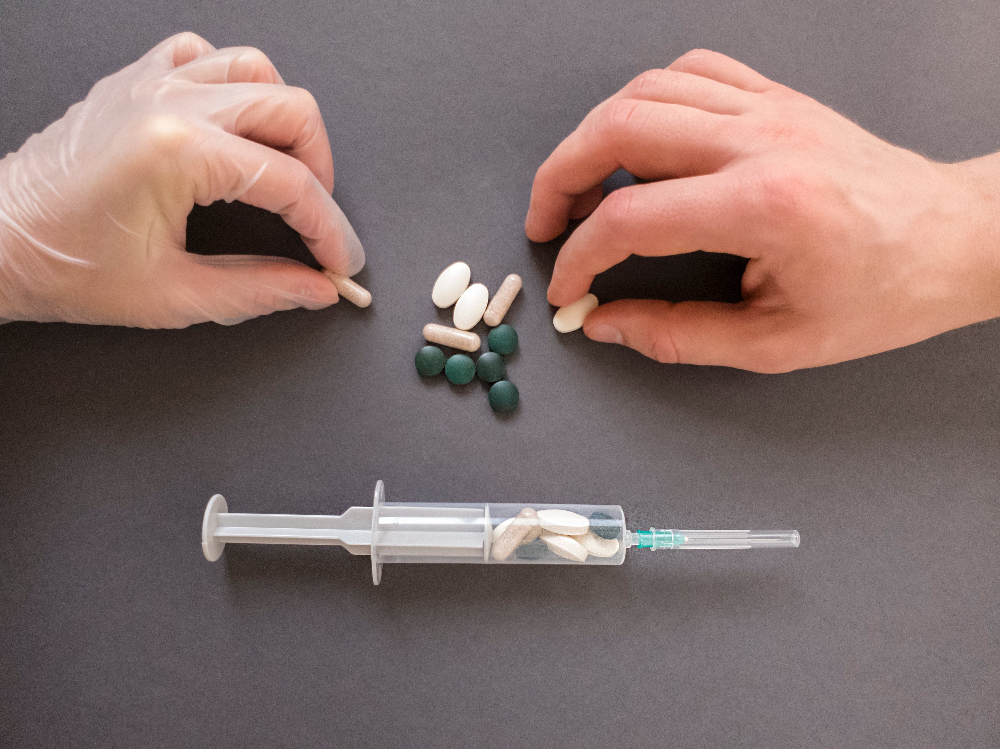 One gloved hand and one ungloved hand hold medicine tablets over a dark table, syringe filled with the tablets sits beside them