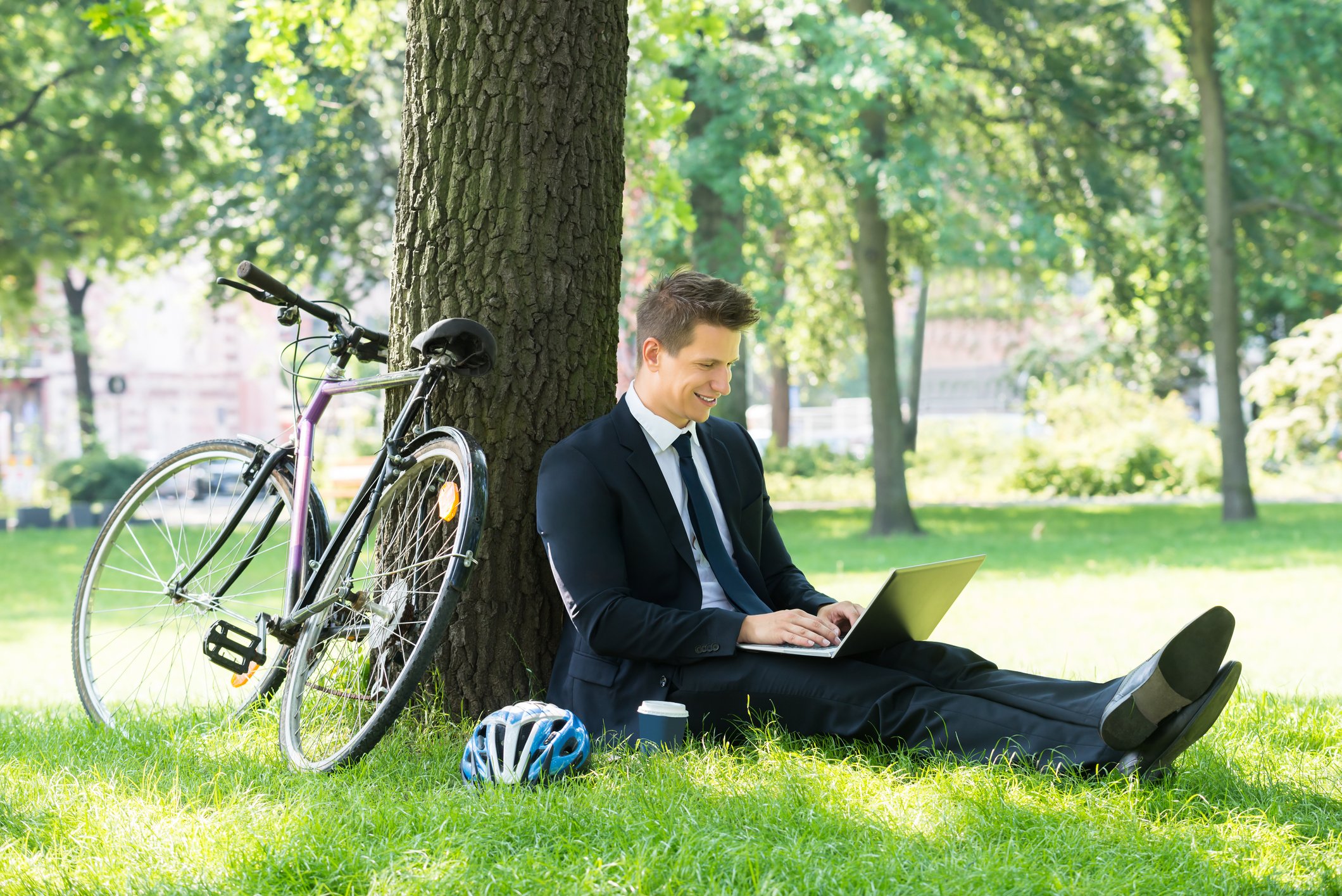 Businessman in casual jacket and tie, sitting under a tree with a bike, working on his laptop.