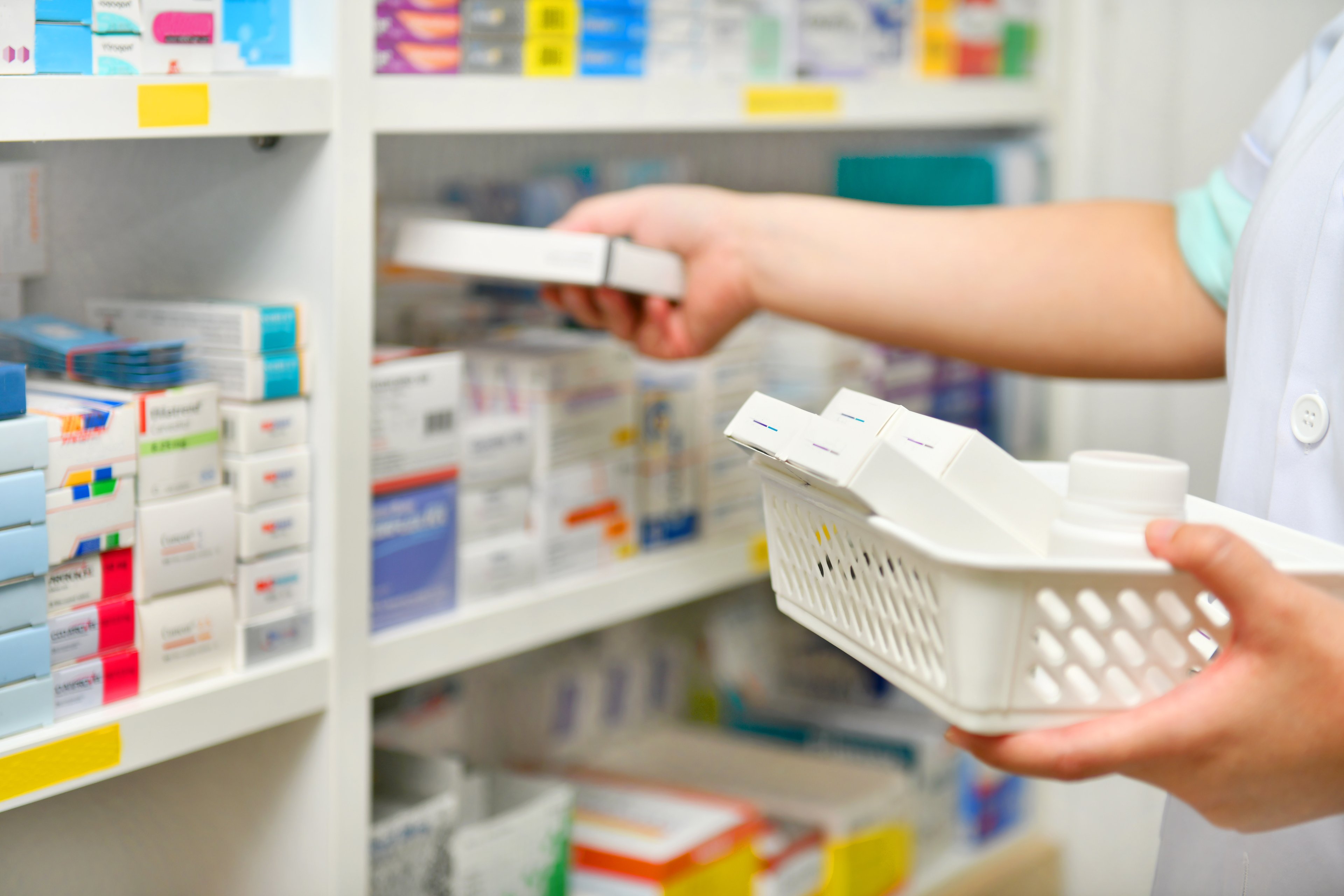 Pharmacist removes medicine package from basket and reaches to place it on shelf alongside other medications.