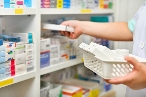 Pharmacist removes medicine package from basket and reaches to place it on shelf alongside other medications.