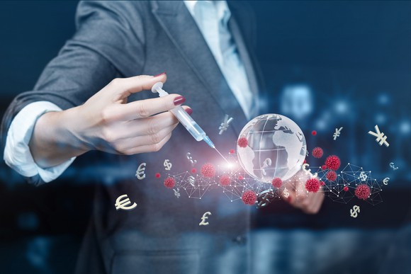 Person holding a syringe up to a small glass globe surrounded by coronavirus cells and currency symbols. 