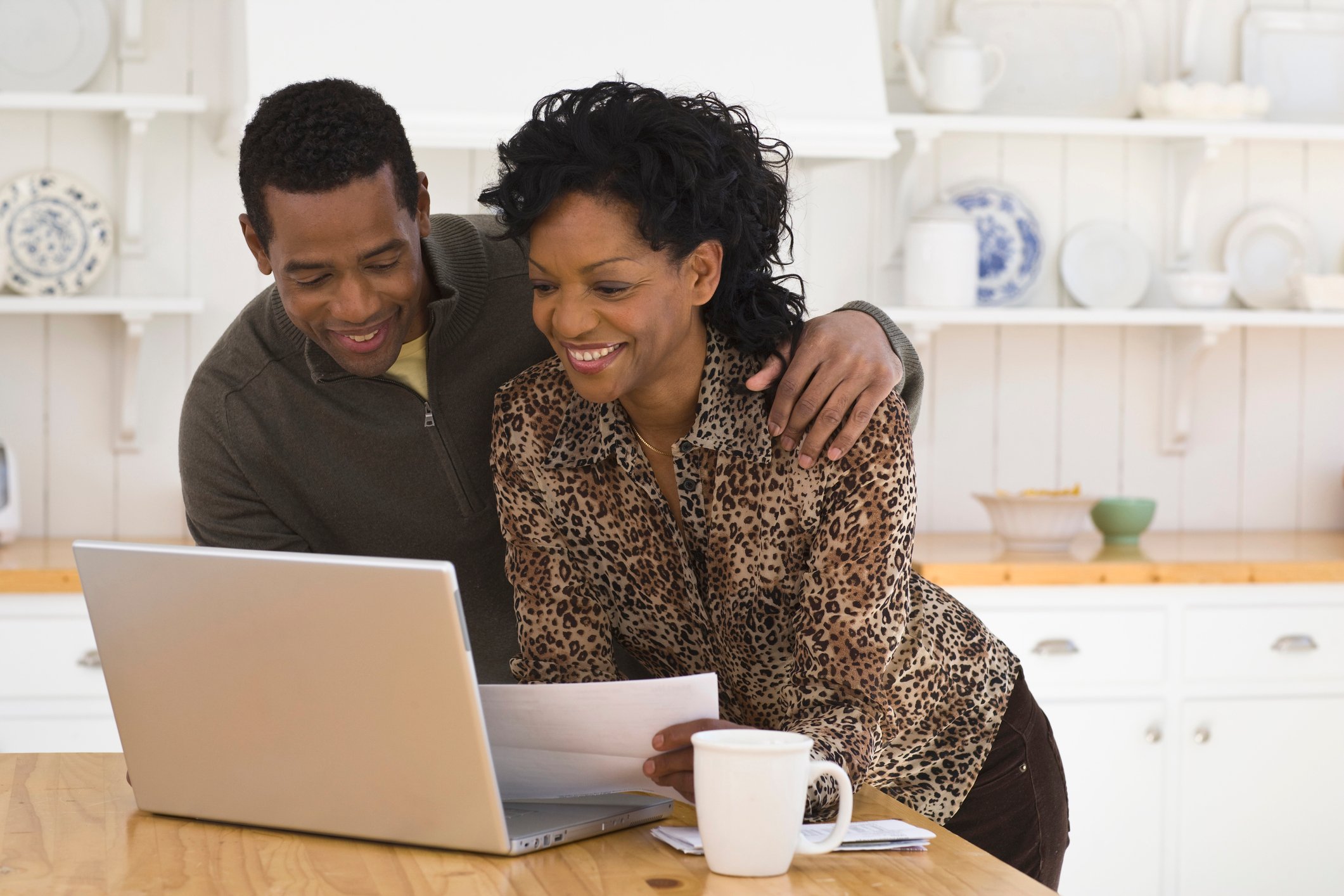 couple holding a piece of paper and looking at a laptop computer screen. 