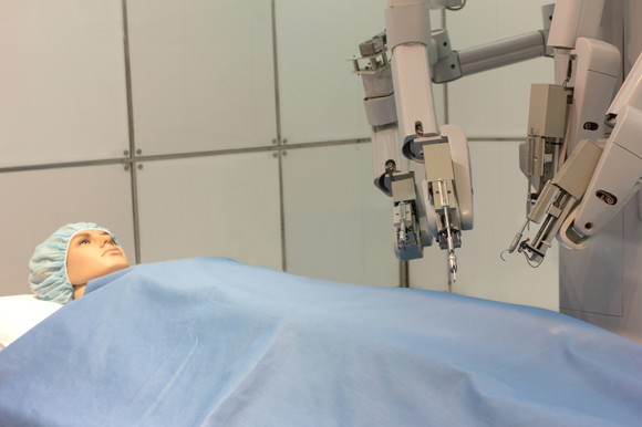 a dummy human on an operating table, covered with a blue sheet and robotic arms positioned above it as if to begin surgery.