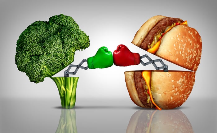 A cheeseburger and a broccoli sparring with mechanical boxing gloves on.