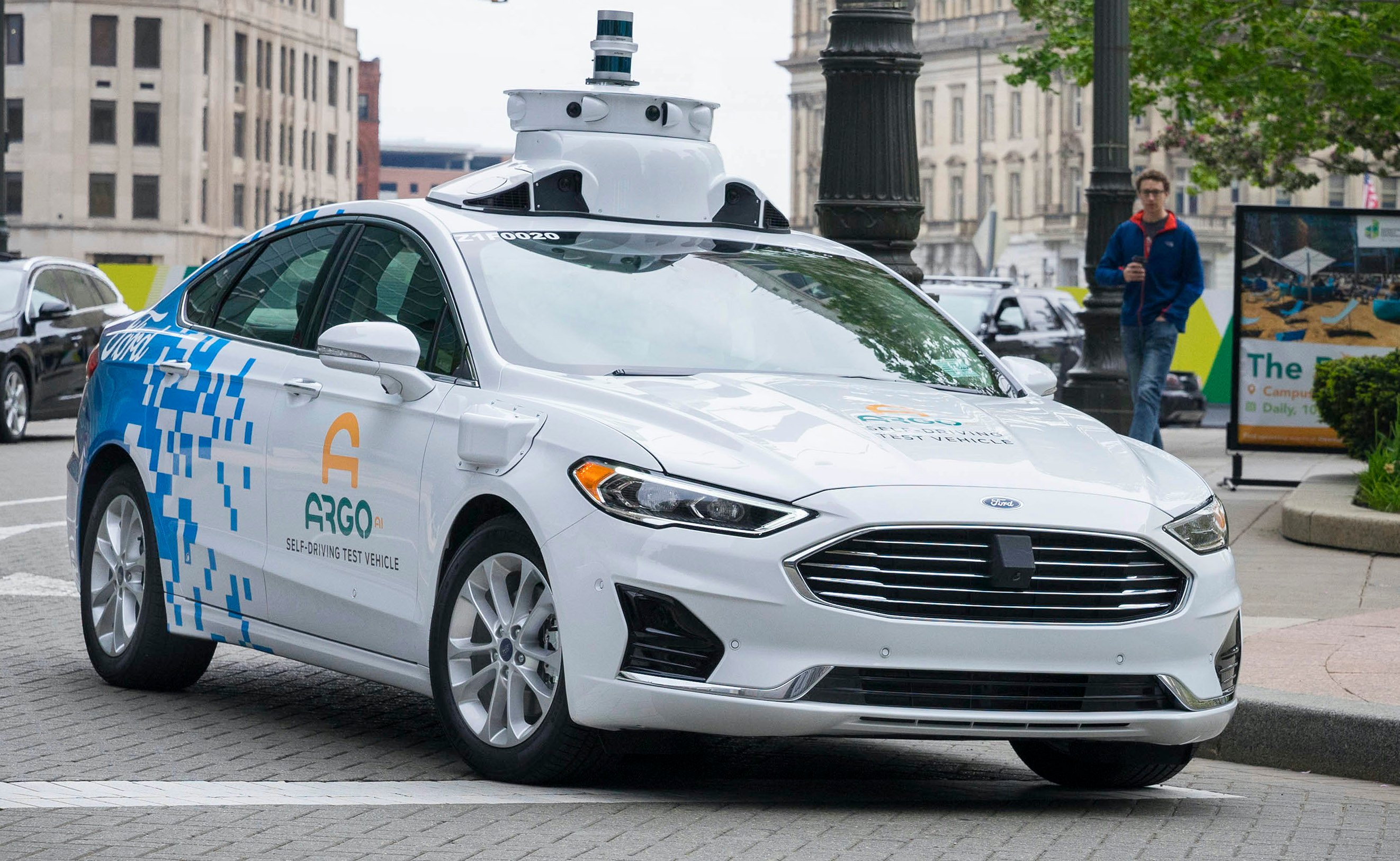 A white Ford Fusion sedan with Argo AI logos and visible self-driving sensor hardware. 