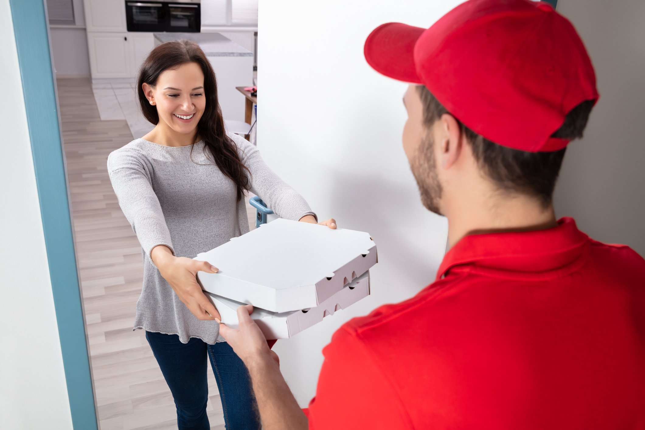 Man in red shirt and red hat delivering two pizzas to a young woman at her door. 