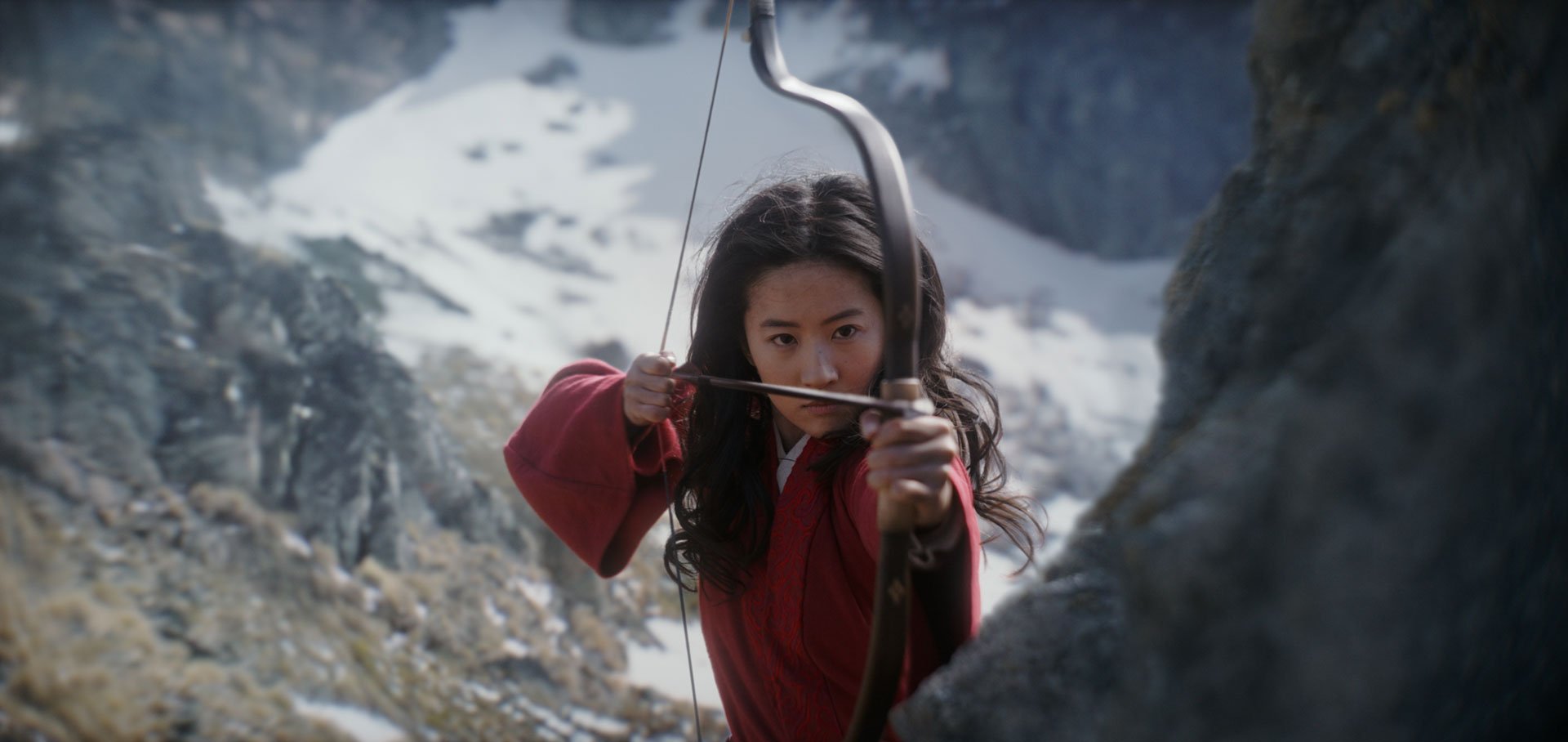 An Asian woman holds a bow and arrow in front of a mountainous background.