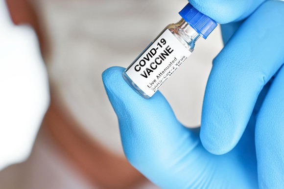 A researcher's gloved hand holds up a vial labeled COVID-19 vaccine.