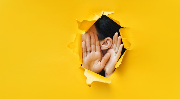 A person trying to hear through a ripped yellow paper wall.
