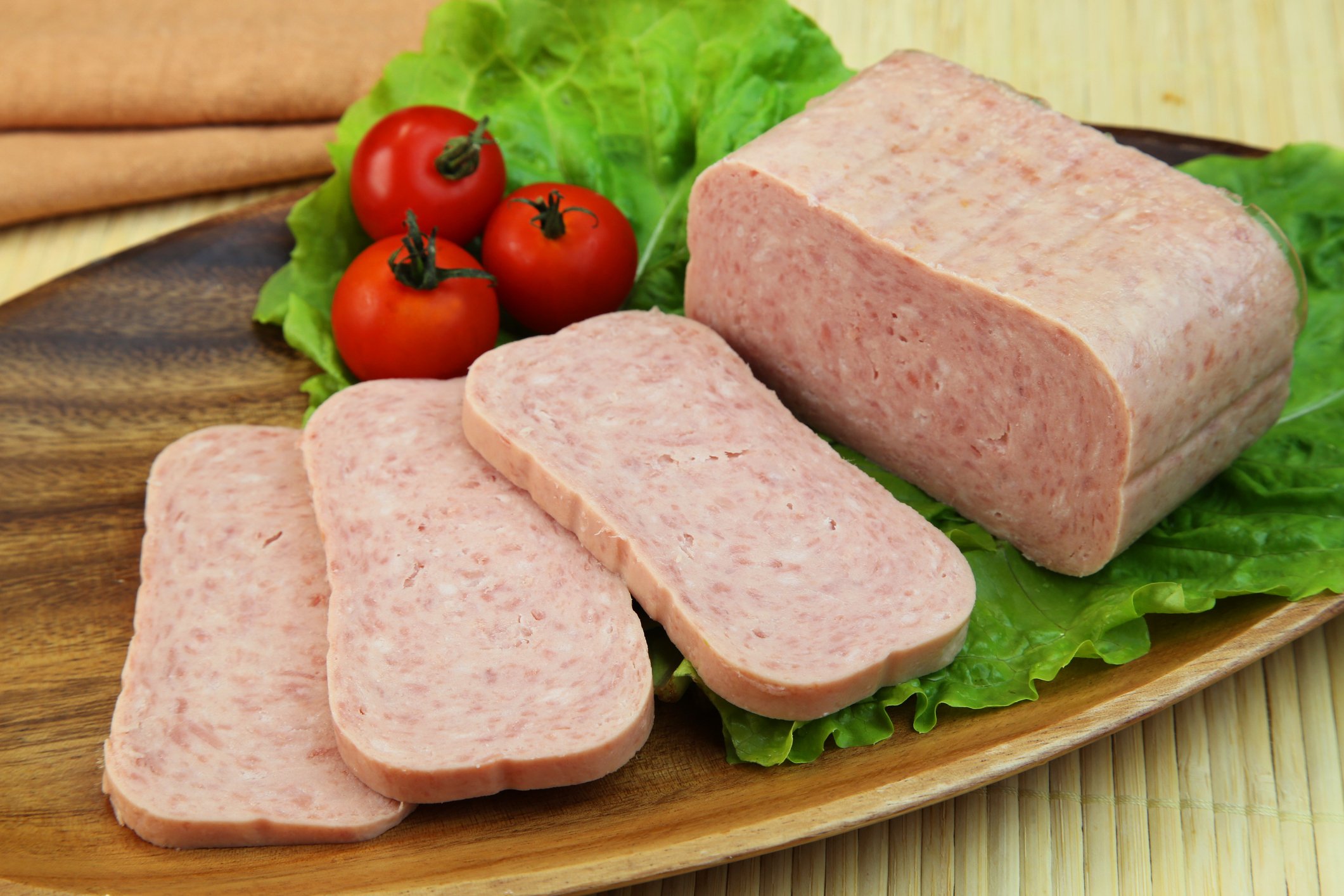 Processed ham similar to spam, sliced on a wooden cutting board with cherry tomatoes and lettuce.