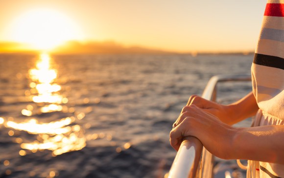 A pair of hands holds a railing on a cruise ship as the sun sets behind it. 
