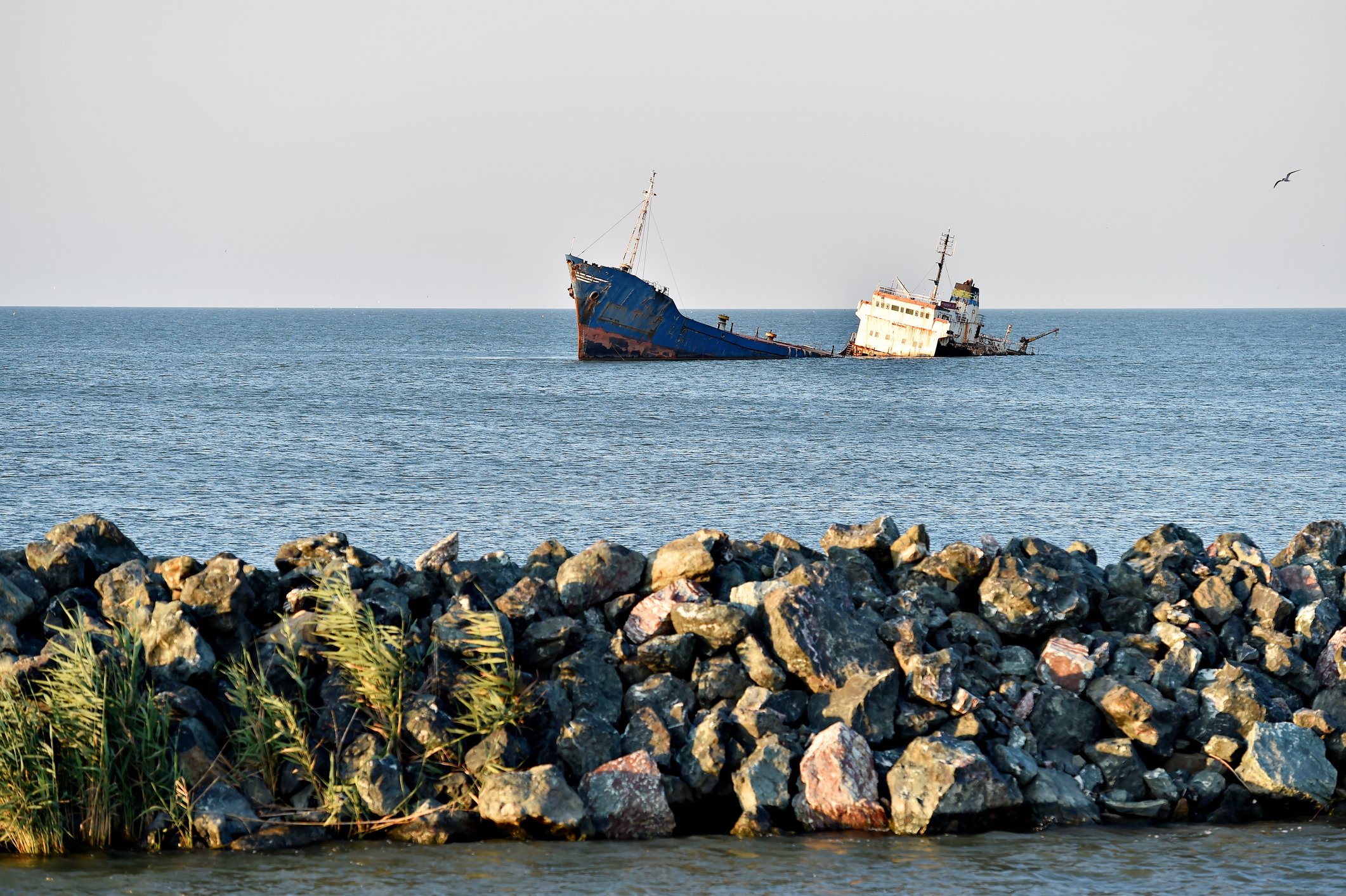 A large modern shipwreck in a sunlit sea with a rocky breakwater in the foreground.