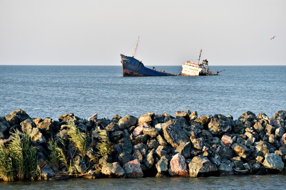A large modern shipwreck in a sunlit sea with a rocky breakwater in the foreground.