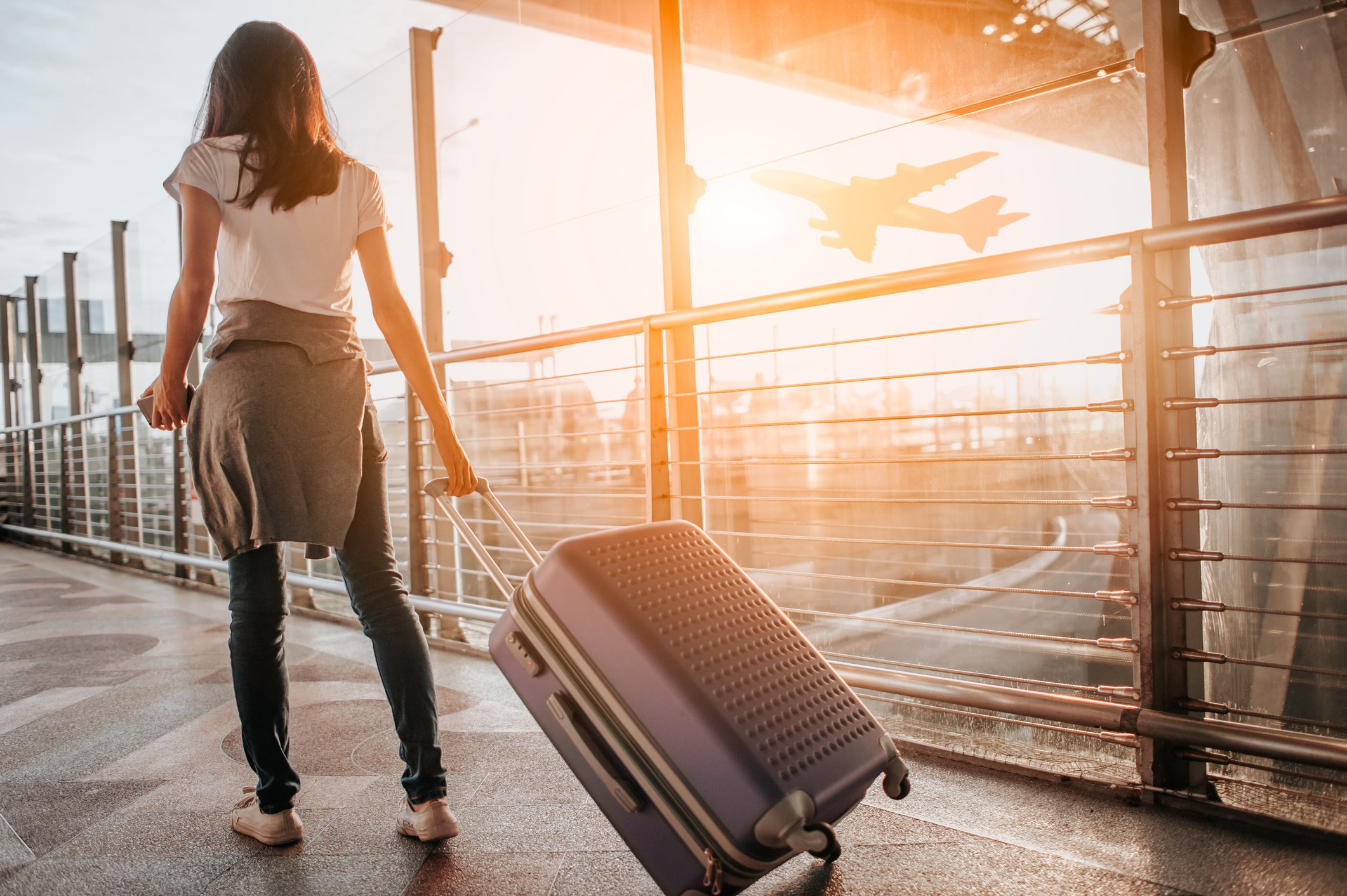 A woman pulls a suitcase through an airport terminal with an airplane taking off in the background. 