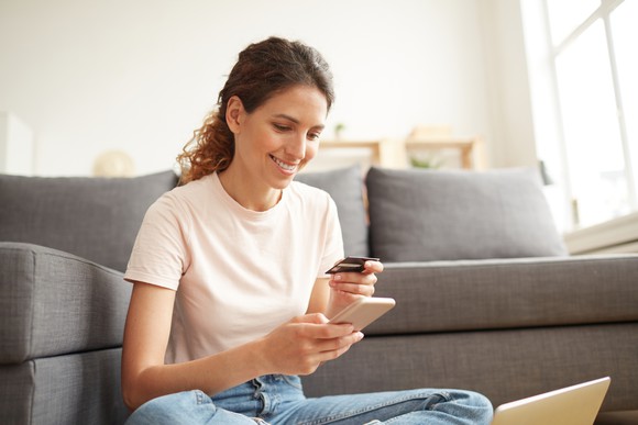 Young woman sitting on the floor as she holds a smartphone in one hand and a credit card in the other.