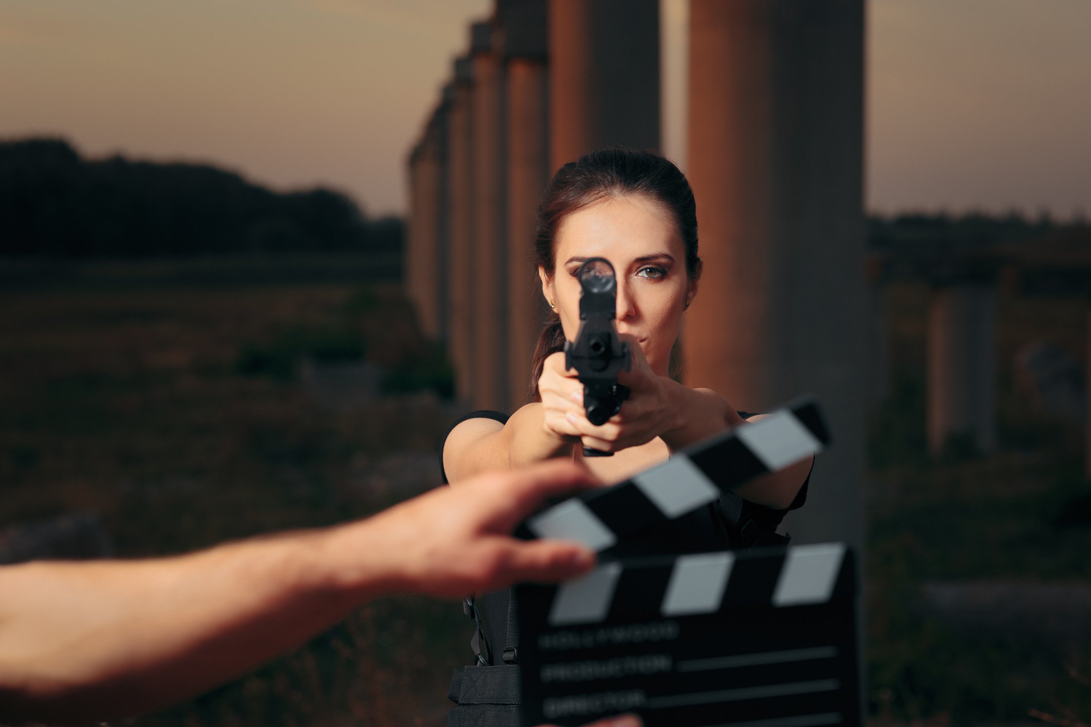 A generic superhero film actress pointing a gun at the camera, a hand holding a clapperboard in the foreground, and pillars in the background.