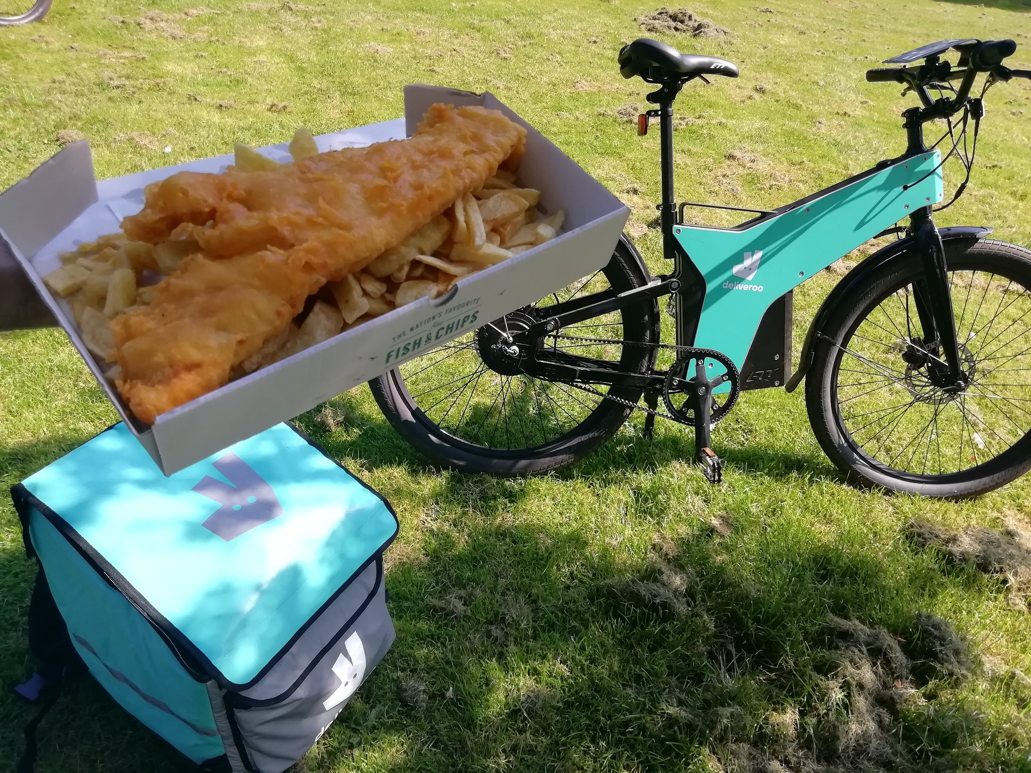 A Deliveroo marked bicycle and delivery pouch resting on grass, with a hand holding a box of fish and chips in the foreground.