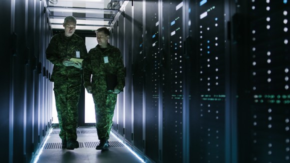 Two military personnel in fatigues walk among a row of servers. 