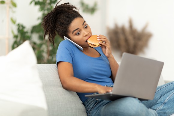 A woman eats a burger while working on her laptop and phone