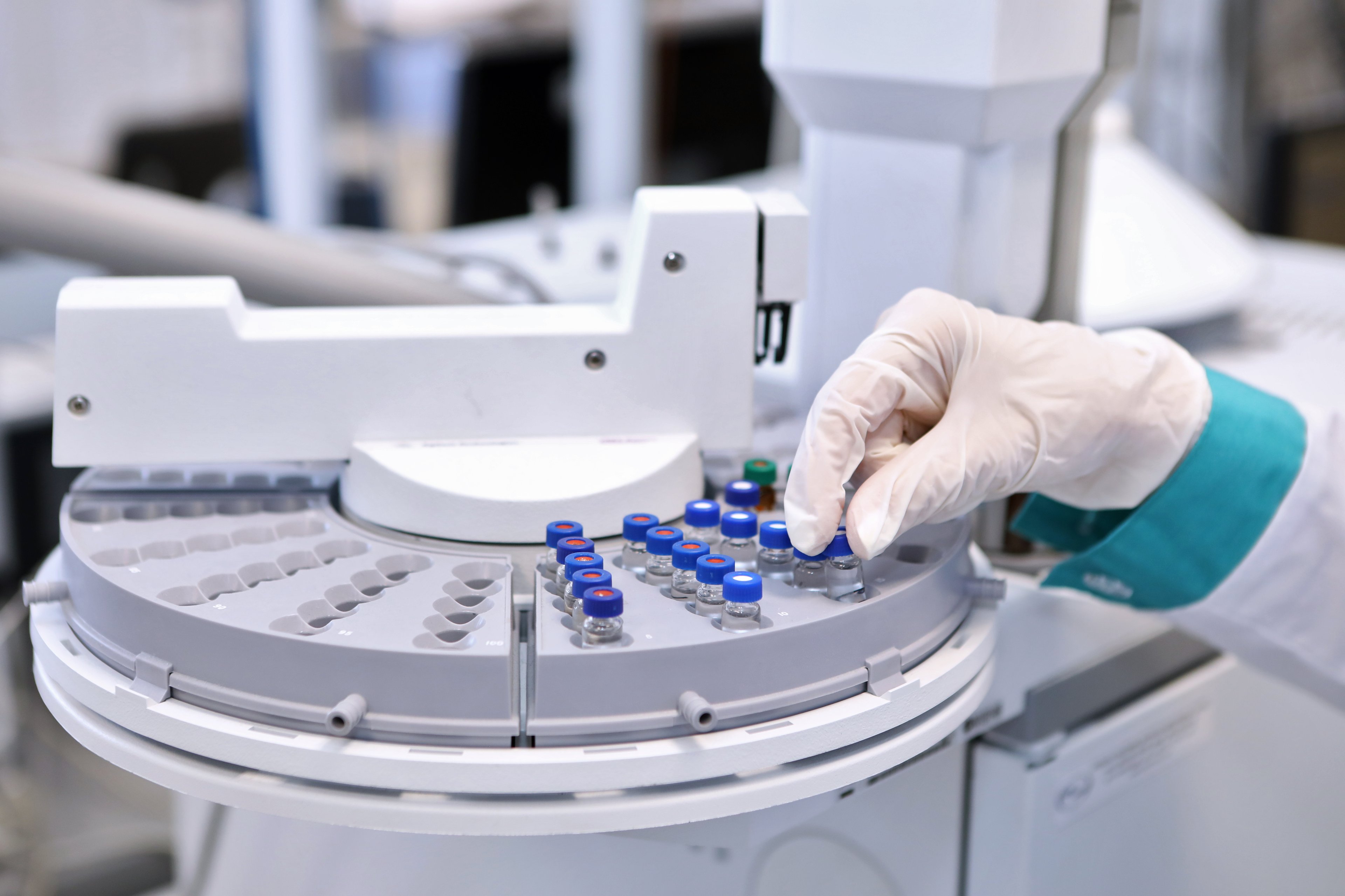 Technician wearing a white coat and glove organizes vials of drug components over a machine.