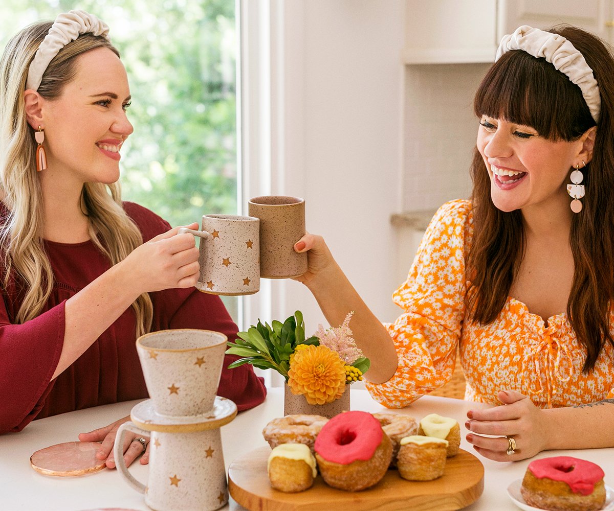 Two smiling women at a kitchen table clinking retro coffee mugs.