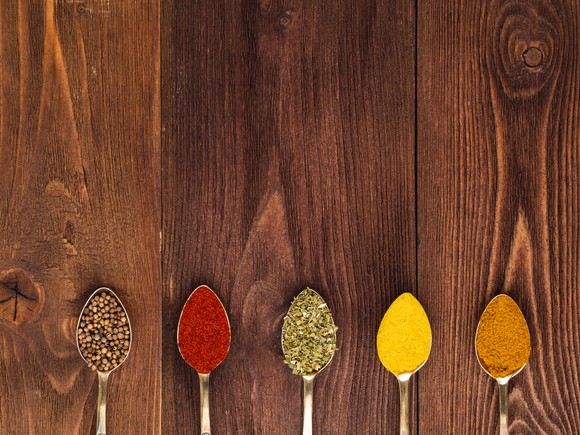 Five spoons filled with colorful spices rest on a wooden table.
