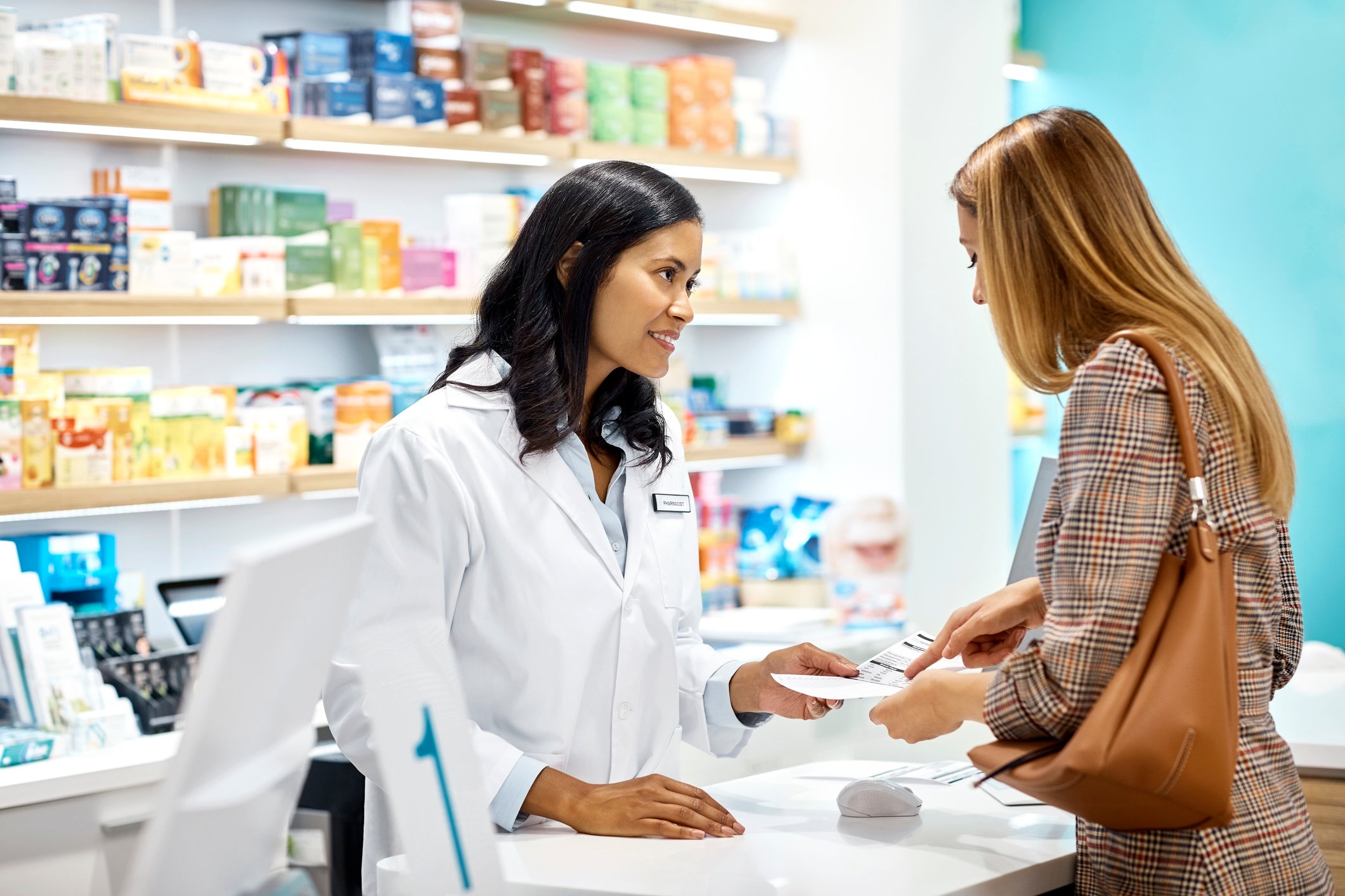 A pharmacist standing in front of shelves of prescriptions, helping a woman at the counter.