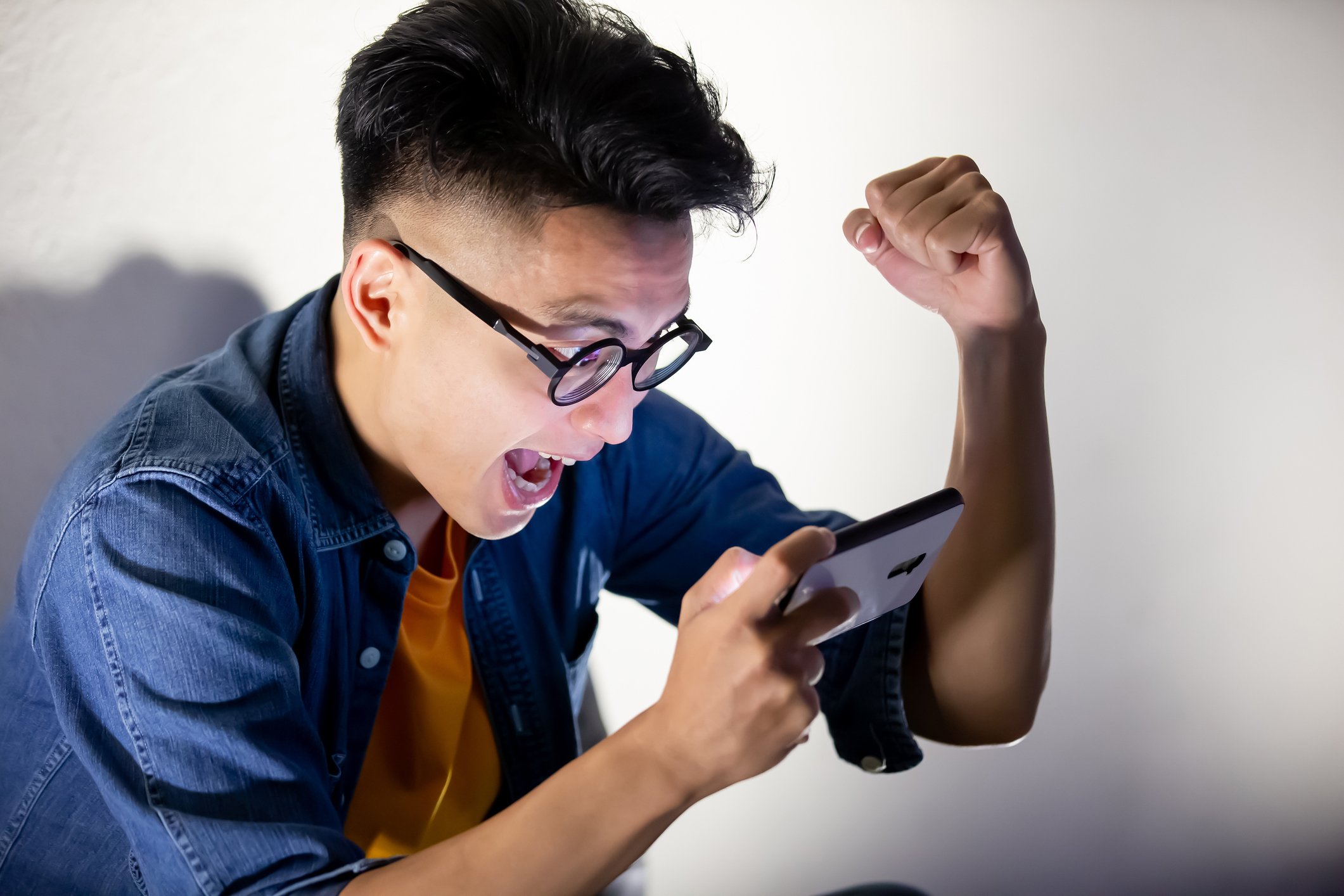 Young man with glasses playing a game on his phone as he raises his left fist in triumph.