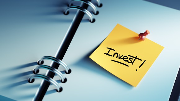 A post-It note saying "invest!" is pinned to the pages of a spiral notebook.