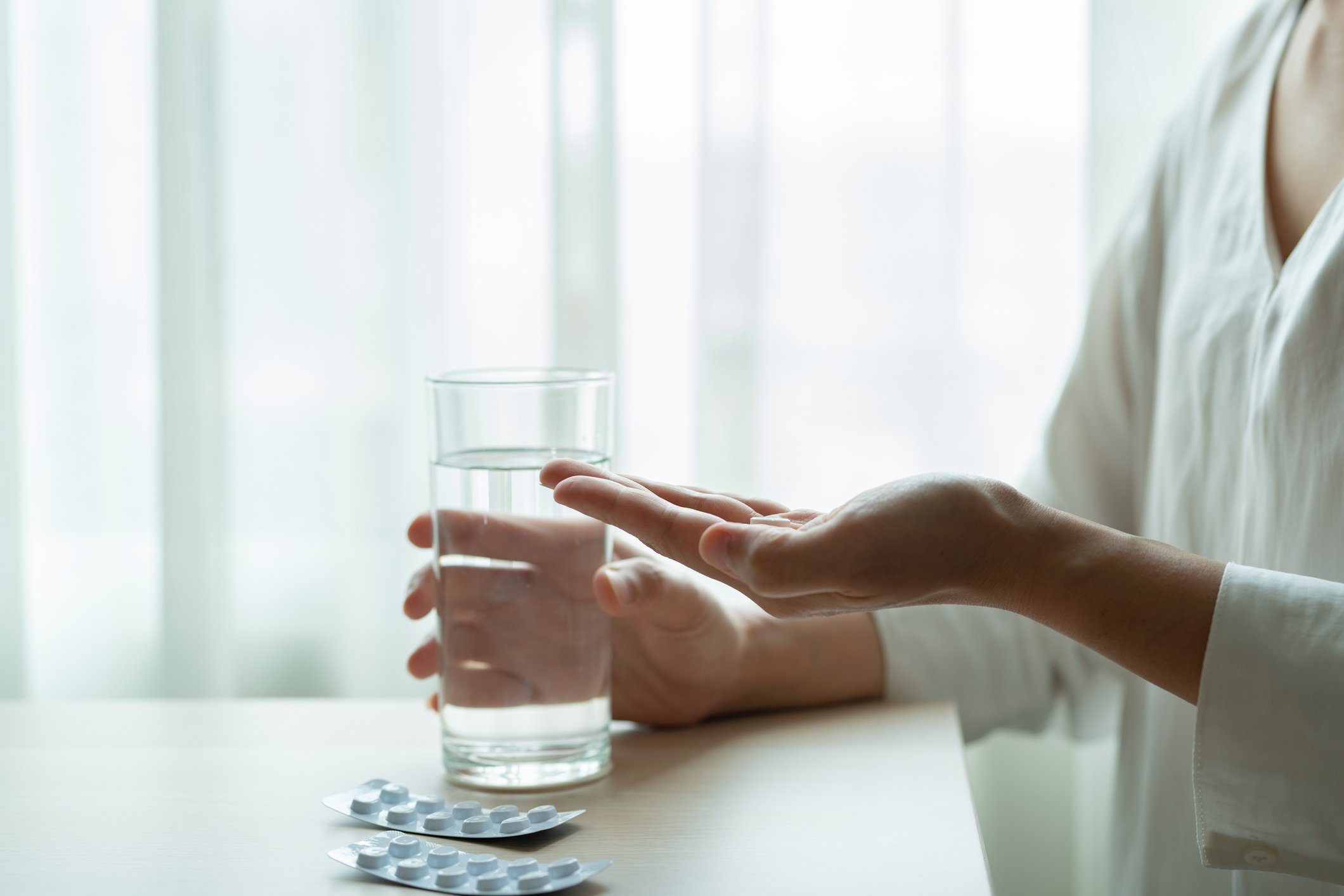 A person at a table with a pack of pills and one in her hand with a glass of water in the other hand.