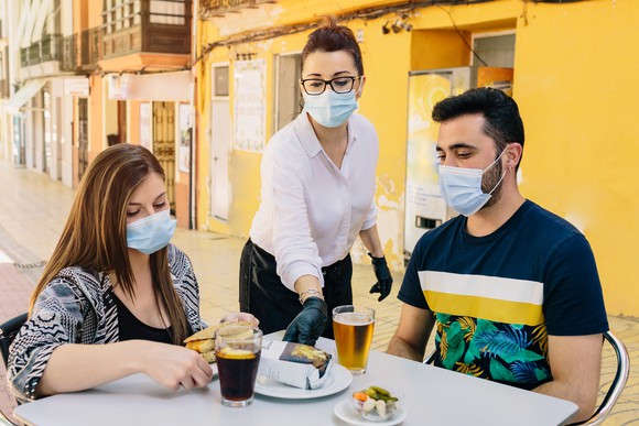 A waitress serves people at an outdoor restaurant while all wear masks.