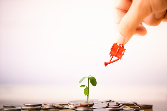 Small watering can watering small plant sprouting from a pile of coins
