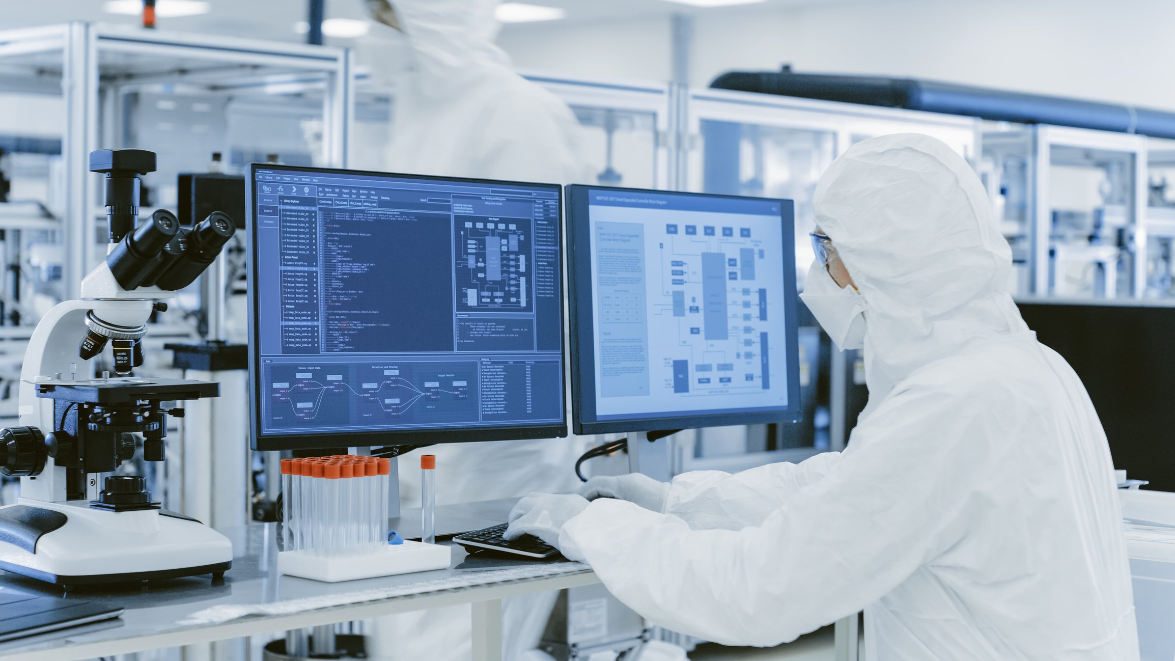 A scientist wearing full protective gear consults a readout from two computer screens.