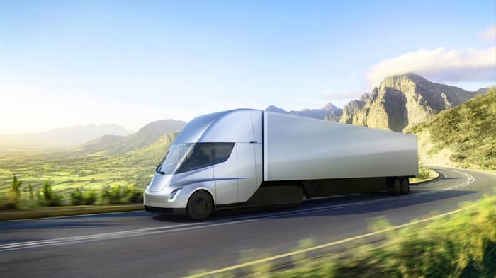 Tesla Semi truck in silver, driving on a road in a grassy mountain meadow.