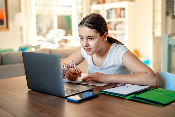 A girl looking a laptop and writing in a notebook.
