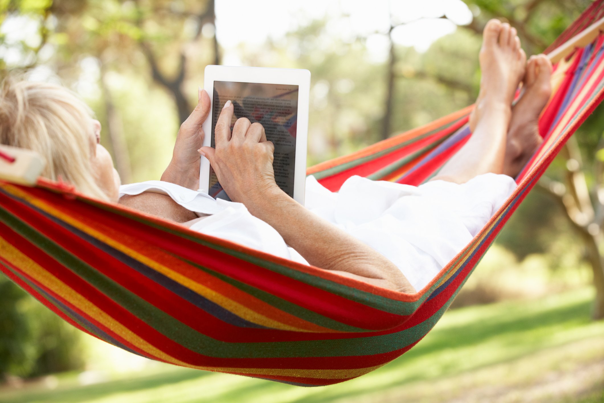 A senior woman relaxes in a hammock while reading an e-book.