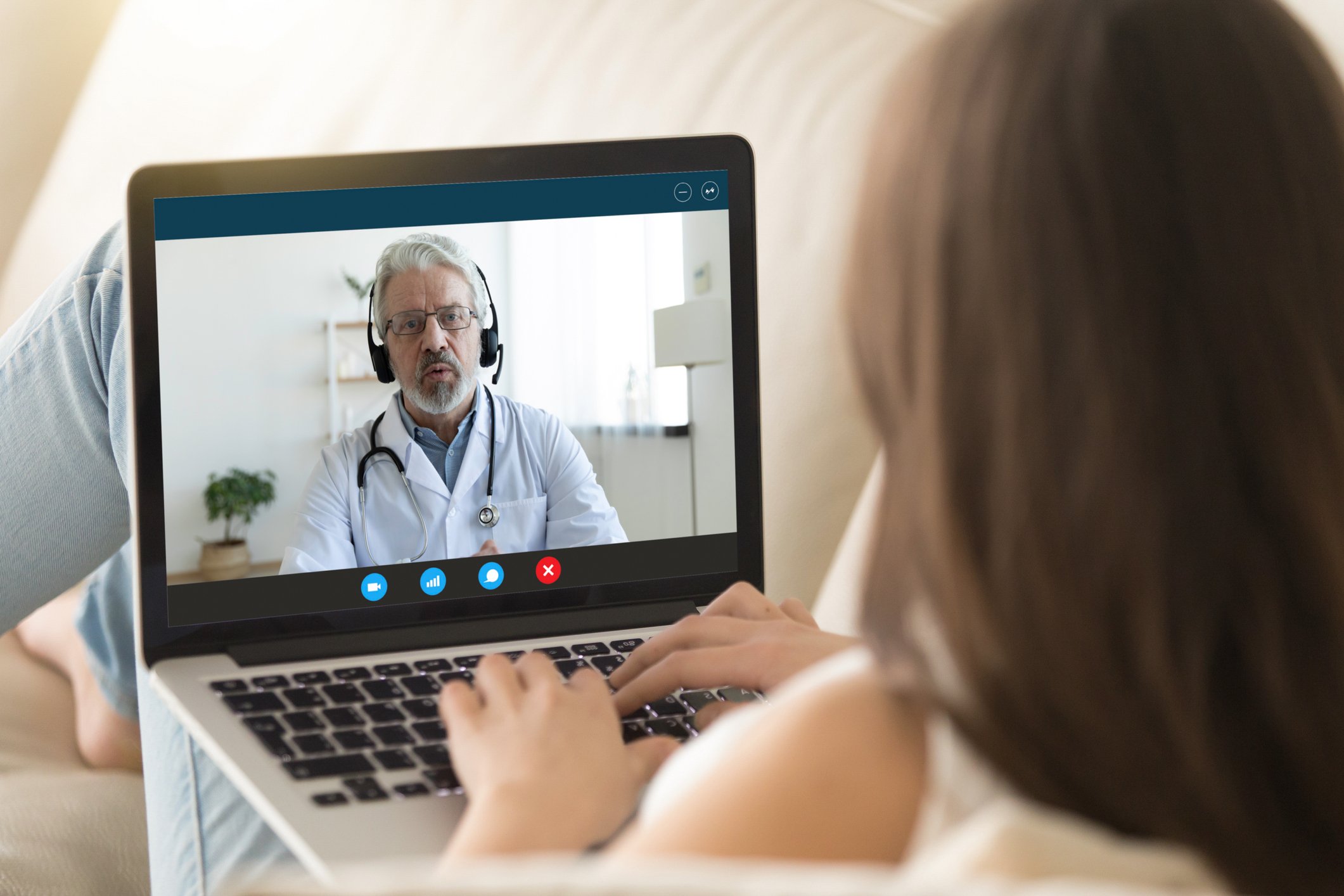 A doctor on the computer screen wearing headphones talks to a woman holding the computer on her lap while sitting on a sofa.