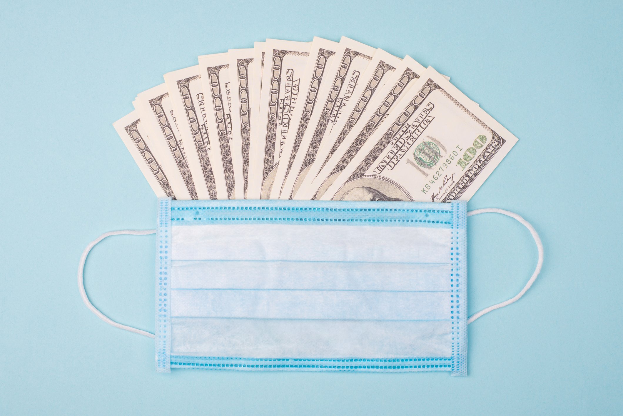 Several hundred dollar bills fanned out with a coronavirus mask over them.
