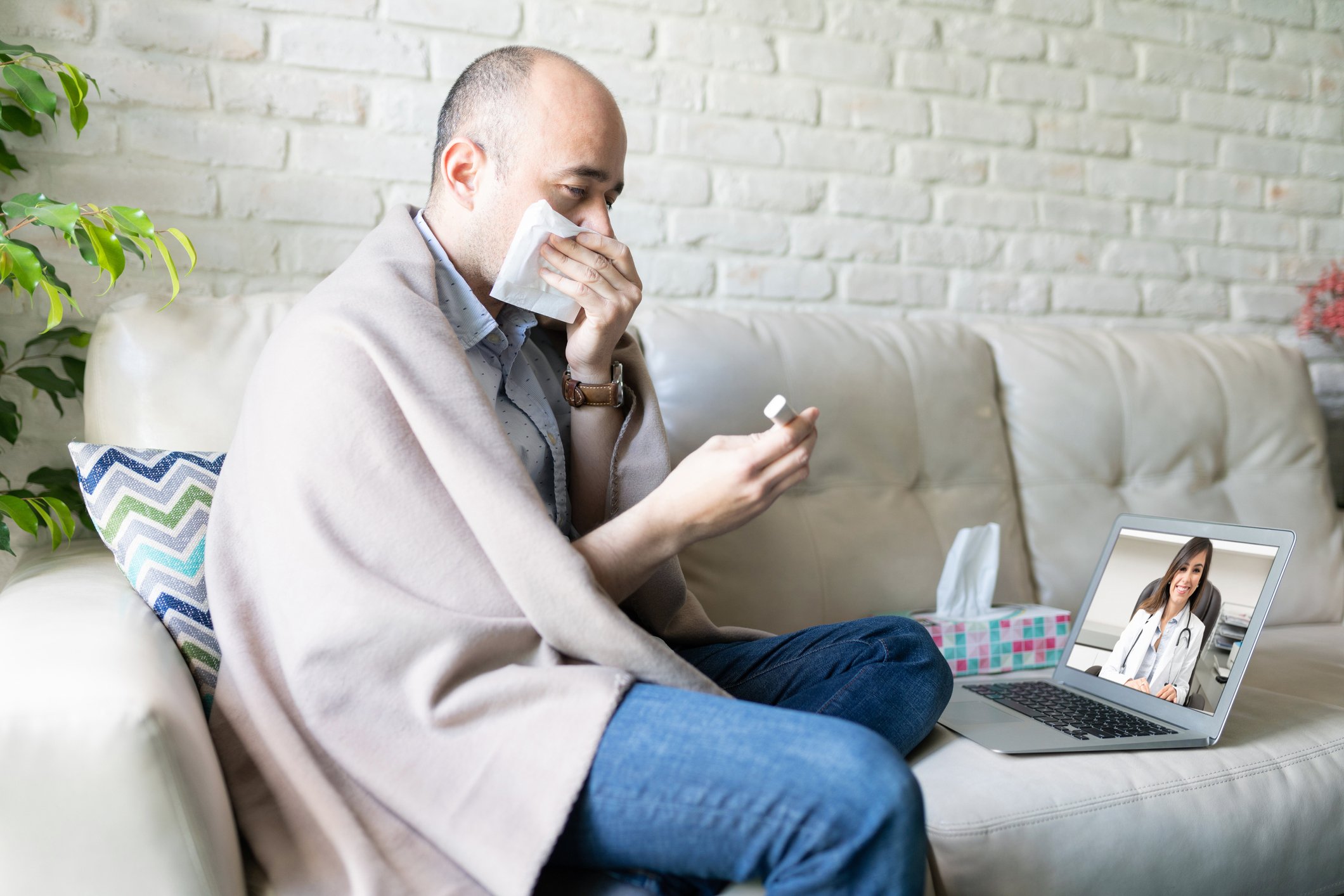 a man covering his mouth with a blanket on his shoulders doing a televisit with a doctor on his sofa