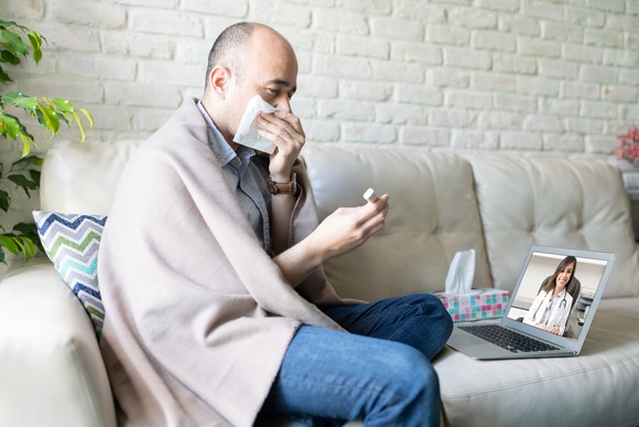 a man covering his mouth with a blanket on his shoulders doing a televisit with a doctor on his sofa