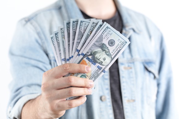 A man wearing a denim shirt holds out a handful of $100 bills.