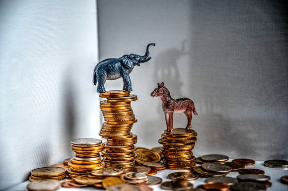 An elephant figurine on a coin stack towering over a donkey figurine on a coin stack.