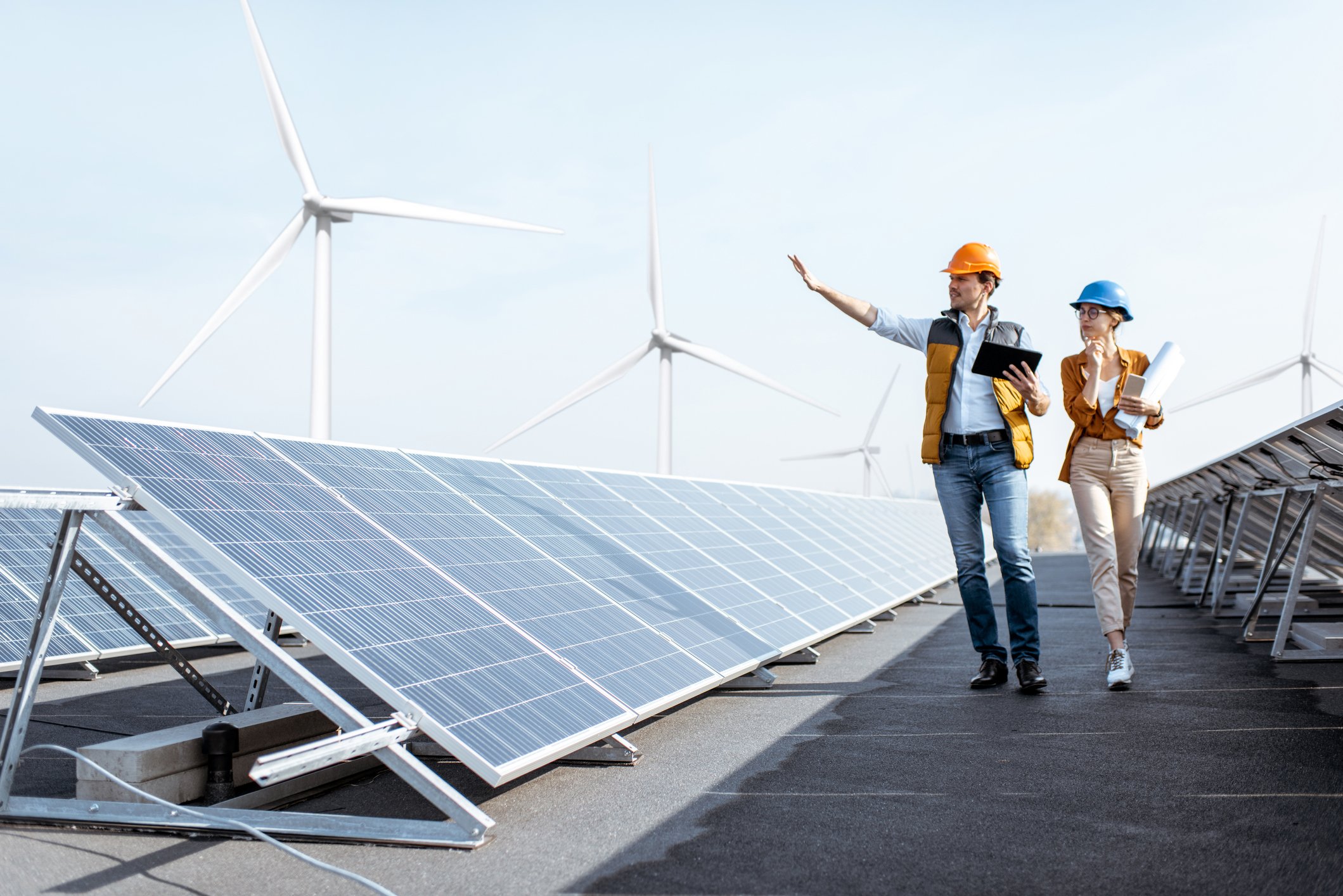 Two workers walk past a solar plant with wind turbines in the background