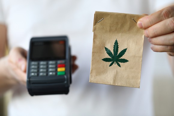 Person holding small paper bag with cannabis leaf on front and credit card processing device 