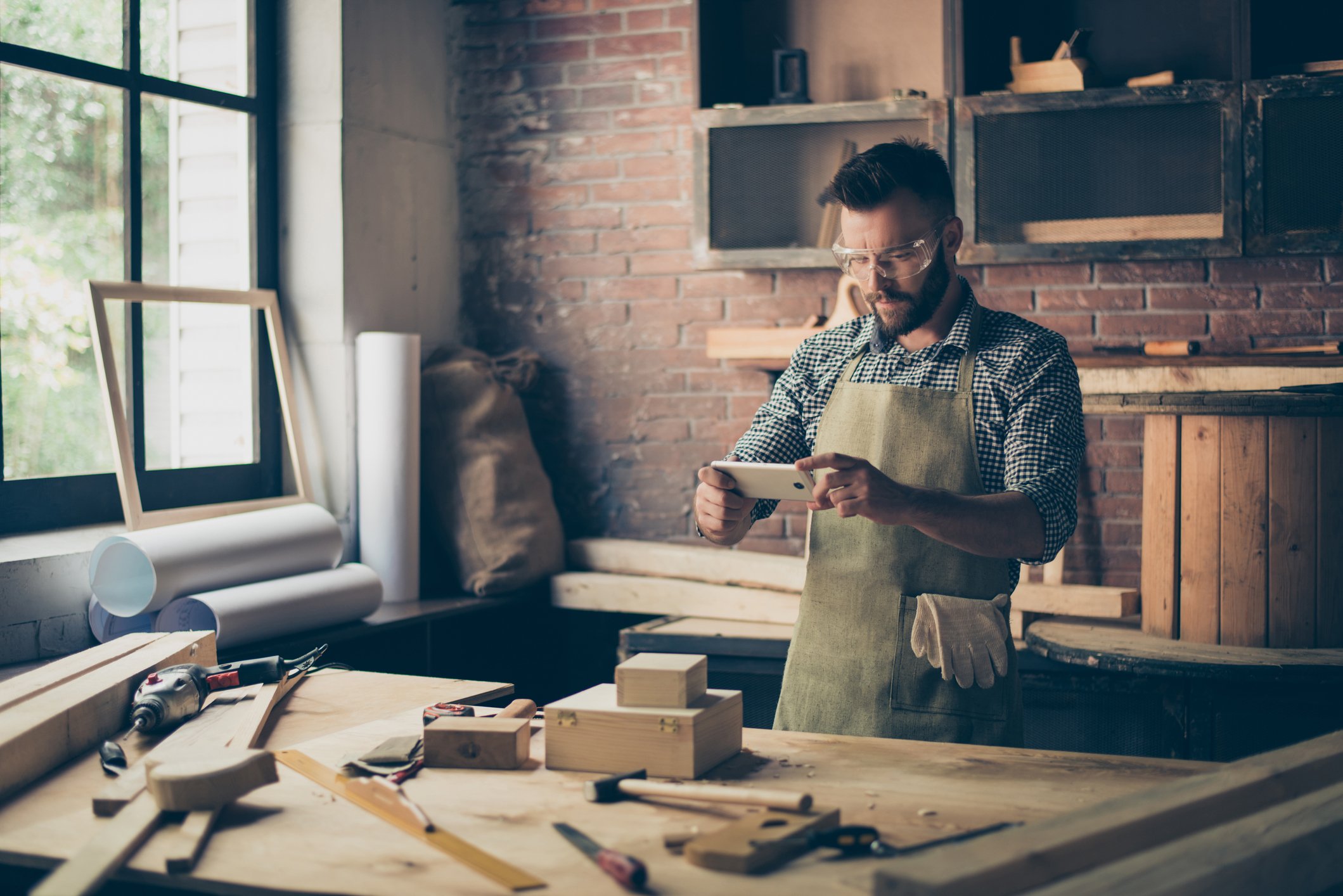 Man with overalls and safety glasses in his workshop using his phone to take a picture of his work.