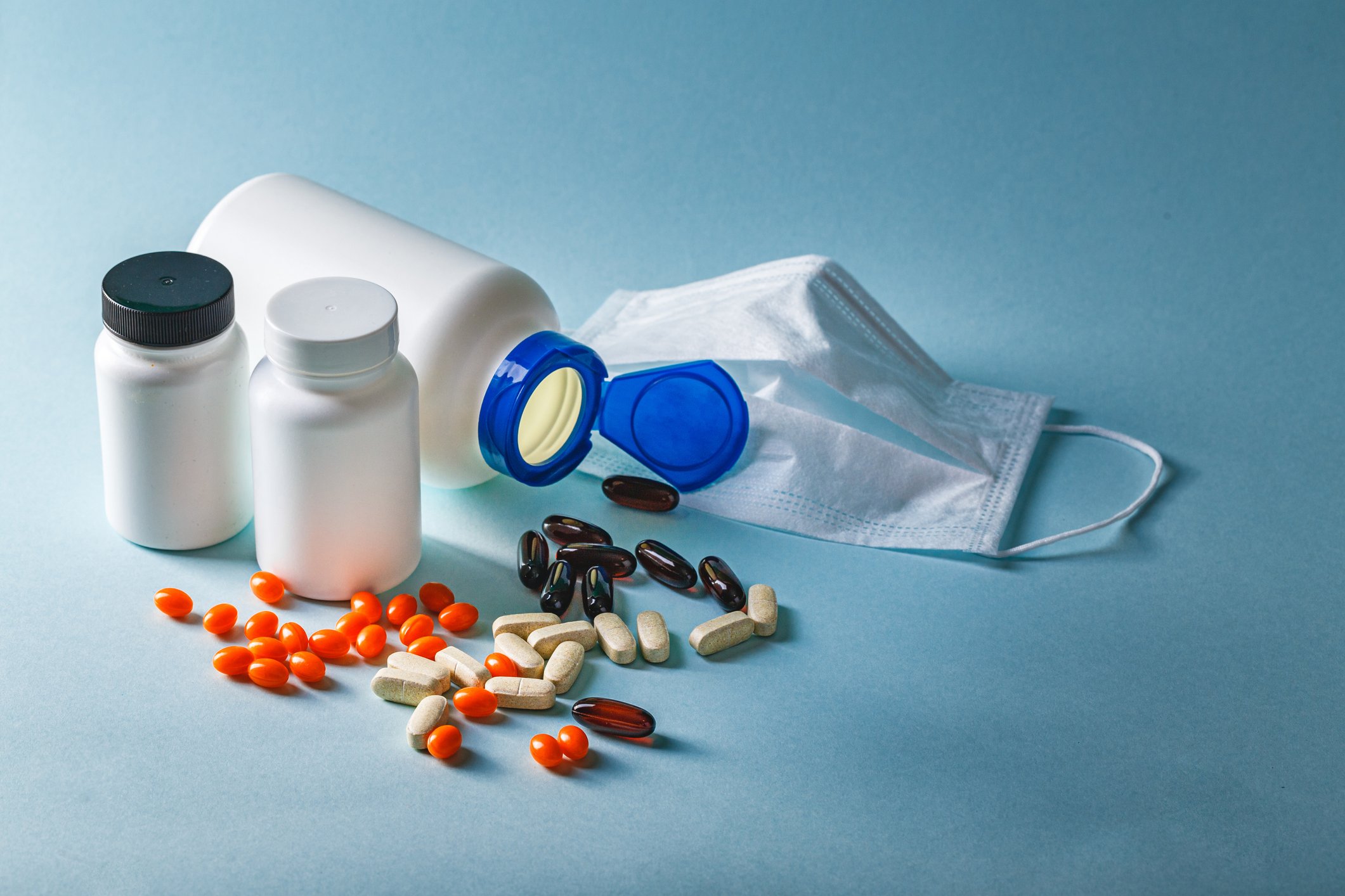 Various colors of pills are scattered in front of three pill bottles, with a surgical mask in the background.